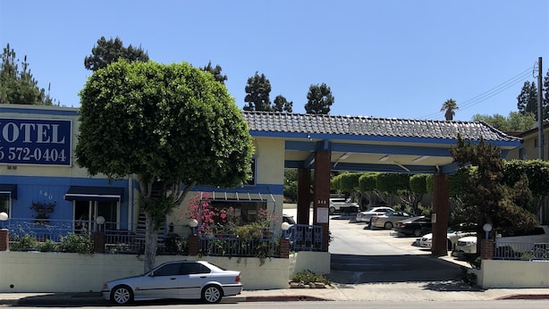 Un hotel con fachada azul, una zona de estacionamiento techada y un árbol en frente.