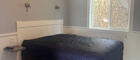 Une chambre à coucher avec un lit, une fenêtre, une table de chevet et un luminaire fixé au mur.