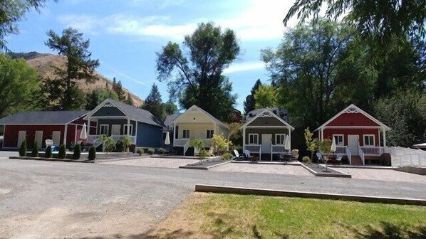 Cozy cottages with pitched roofs and porches line a lush lawn amidst a hilly landscape.