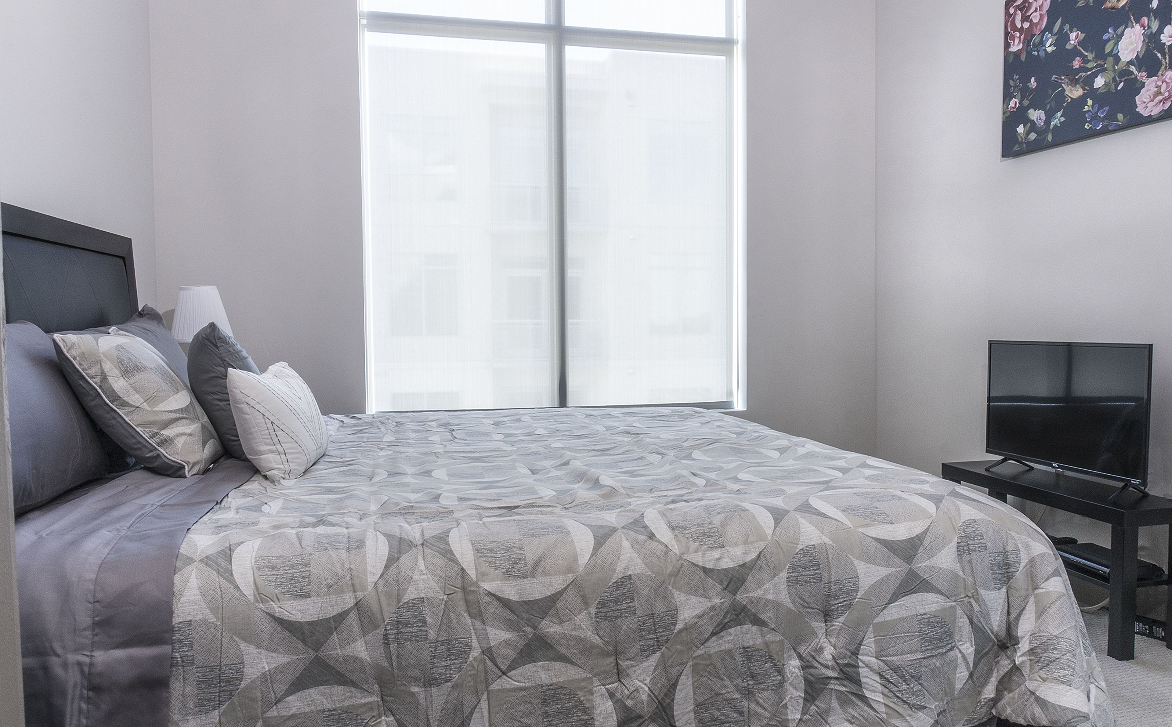 A modern bedroom with a large bed, a TV on a stand, and a window with blinds.