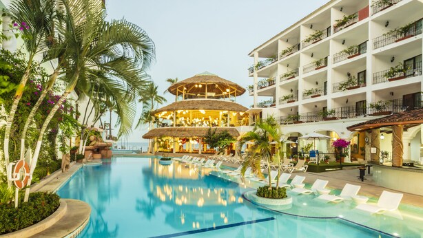 Et resort med poolområde, en stråtækt bygning, liggestole og en bygning med mange etager og altaner.