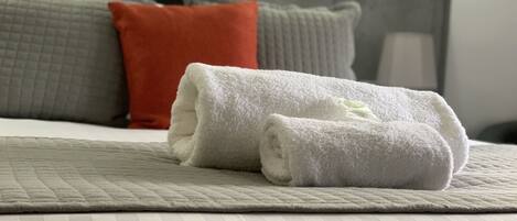 A neatly made bed with a rolled white towel, a grey quilted blanket, and a grey pillow with a red accent.