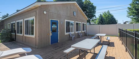 Un edificio de madera con puerta azul, ventanas grandes y mesas de picnic en una terraza de madera.