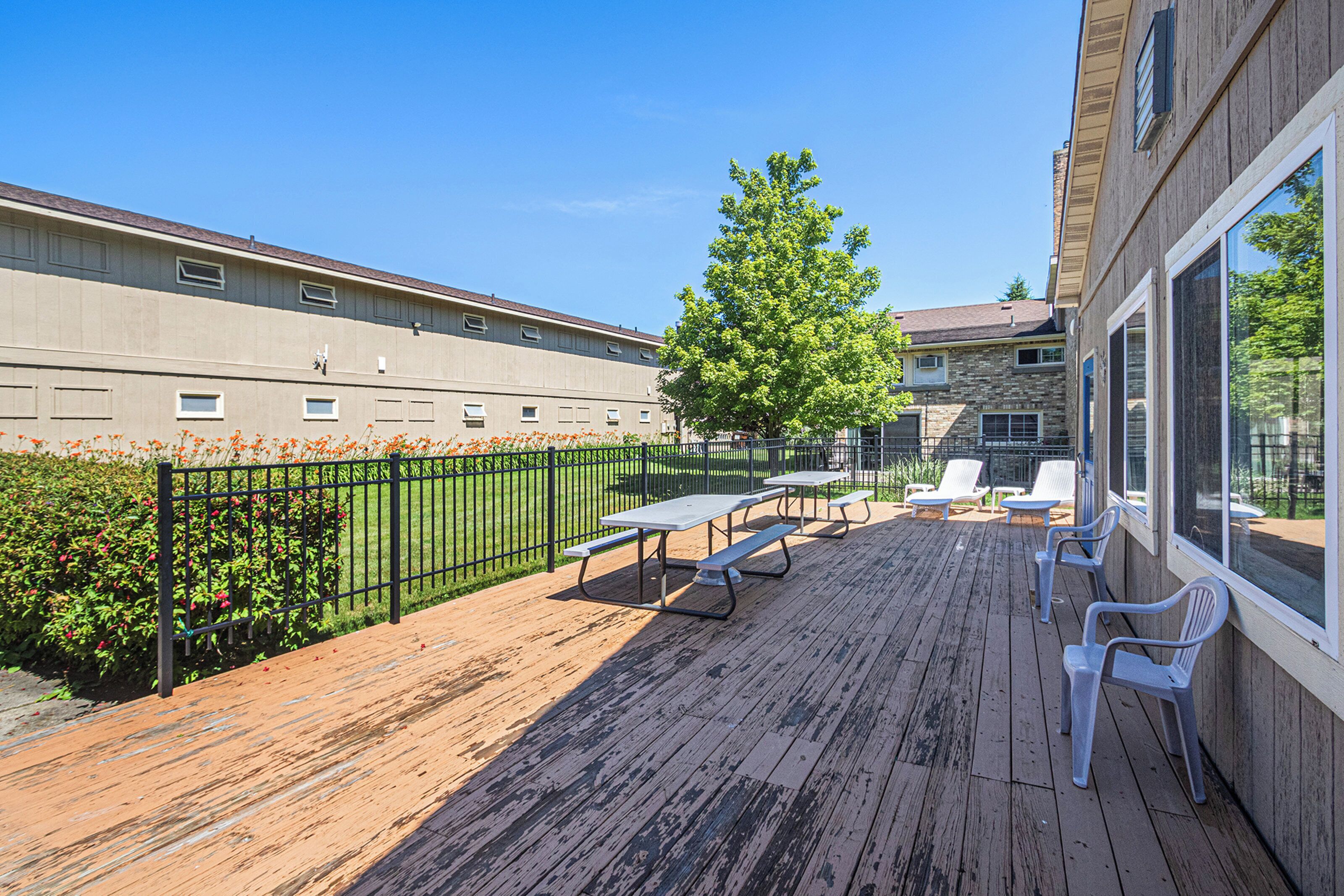 An outdoor deck with picnic tables and chairs, a fenced garden area, and a building in the background.