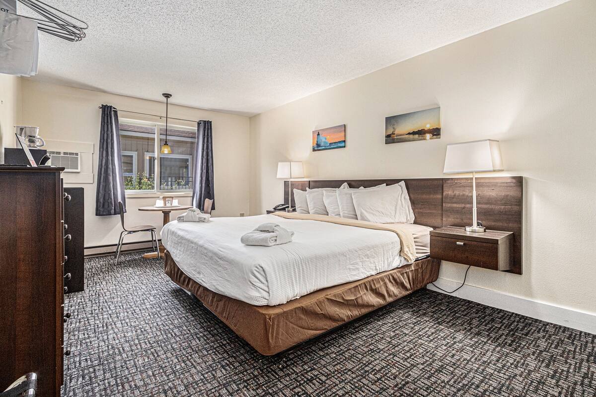 A hotel room with a large bed, a desk, a chair, a window with curtains, and artwork on the wall.