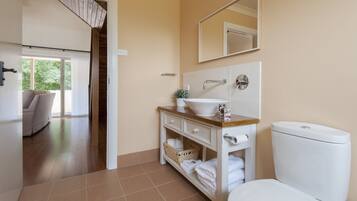 Een badkamer met toilet, wastafel en spiegel. Er is een houten opbergmeubel met lades en een mandje. Een handdoekhouder is zichtbaar en de kamer heeft een betegelde vloer.