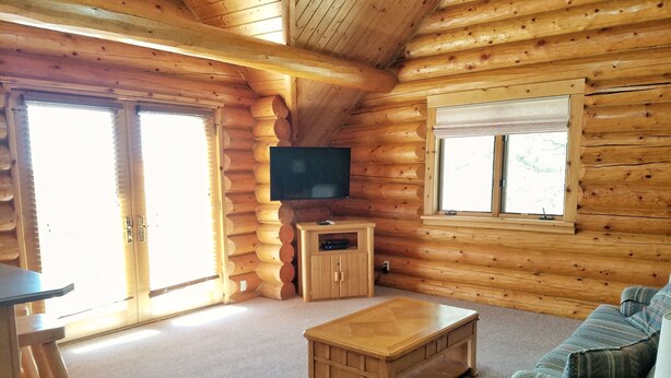 Ein Interieur einer Blockhütte mit einem TV-Schrank aus Holz, einer Couch, einem Couchtisch und einem Fenster mit Jalousien.