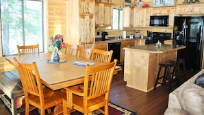 Interior de una cabaña de madera con una mesa de comedor, sillas, una cocina equipada con microondas, estufa y refrigerador, y un ventanal grande.