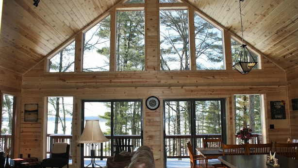 Una cabaña espaciosa con techo alto, ventanas grandes que ofrecen vistas del bosque circundante, un rincón acogedor con sofá y sillones, y una mesa de comedor con sillas.