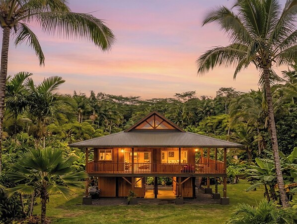 A two-story wooden house with a balcony, surrounded by palm trees and lush greenery.