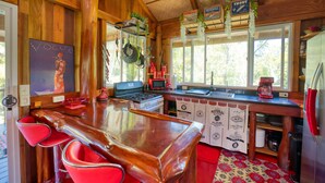 Interno di una baita in legno con angolo bar, sgabelli rossi, una cucina completa di piano cottura e forno a microonde, e piante sospese.