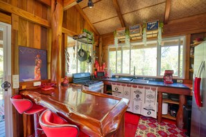 A wooden cabin interior with a bar area, red bar stools, a kitchen with a stove, microwave, and hanging plants.