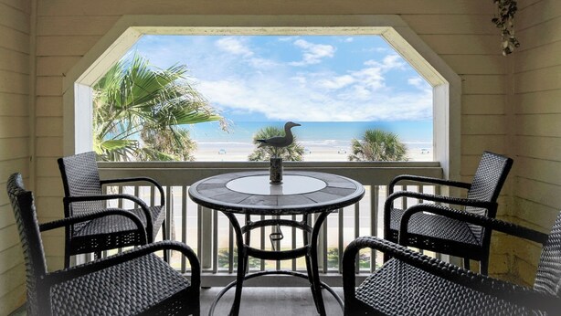 Ein Balkon mit Korbstühlen und einem Tisch, der Blick auf einen Strand mit Palmen und dem Meer bietet.