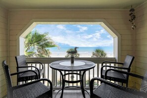 Een balkon met rieten stoelen en een tafel, met uitzicht op een strand met palmbomen en de oceaan.