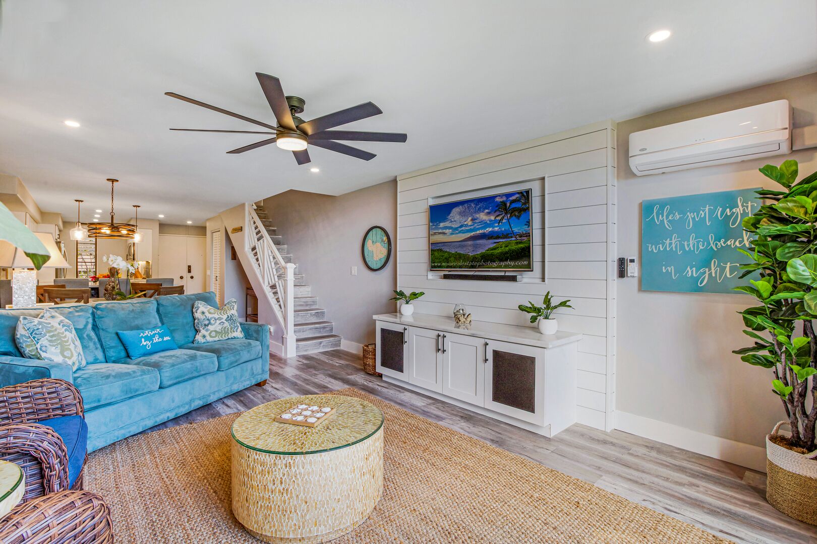 A modern living room with a blue sofa, a glass coffee table, a white cabinet, a ceiling fan, and a painting of a beach scene.