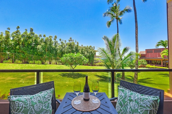 A balcony with a table set for two, featuring a bottle of wine and two glasses. The view includes a well-maintained lawn and palm trees.