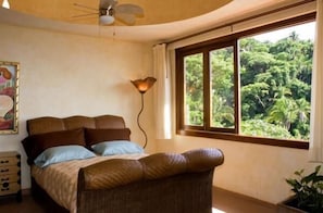 Une chambre à coucher avec un lit, un ventilateur de plafond, une fenêtre donnant sur un paysage verdoyant et une lampe fixée au mur.