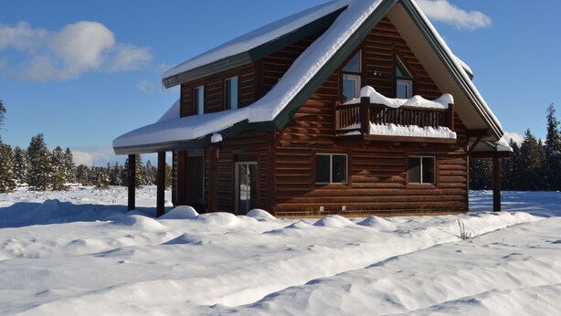 A log cabin with a snow-covered roof and a porch, surrounded by a snowy landscape.