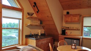 A cozy cabin interior with a dining table, chairs, a kitchen area with wooden cabinets, and a large window offering a view of the outdoors.