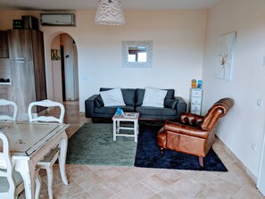 A living room with a sofa, a coffee table, a leather armchair, a dining table with chairs, and a kitchen area in the background.