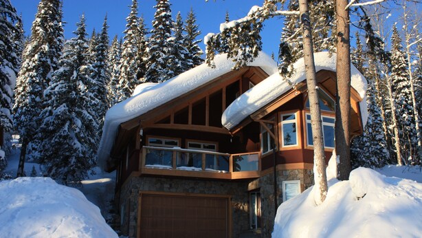 Una cabaña nevada con fachada de piedra, ventanas grandes y un balcón de madera.