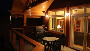 Un patio al aire libre con una terraza de madera, una mesa redonda y sillas. Hay una parrilla y una lámpara cerca de la ventana.
