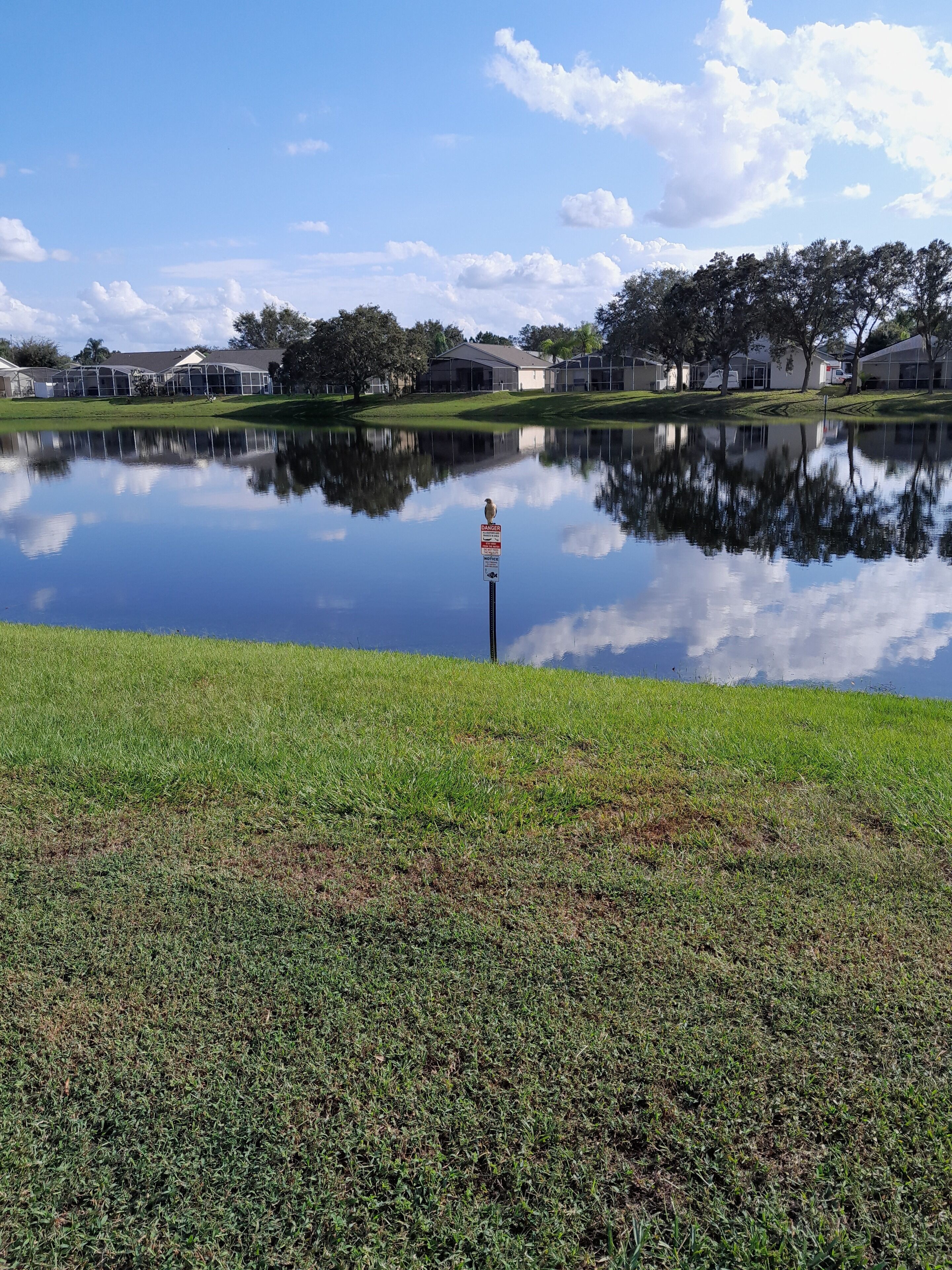 Un lago tranquillo con il riflesso del cielo e degli edifici circostanti. Un cartello con la scritta 'DANGER' è visibile su un palo immerso nell'acqua.
