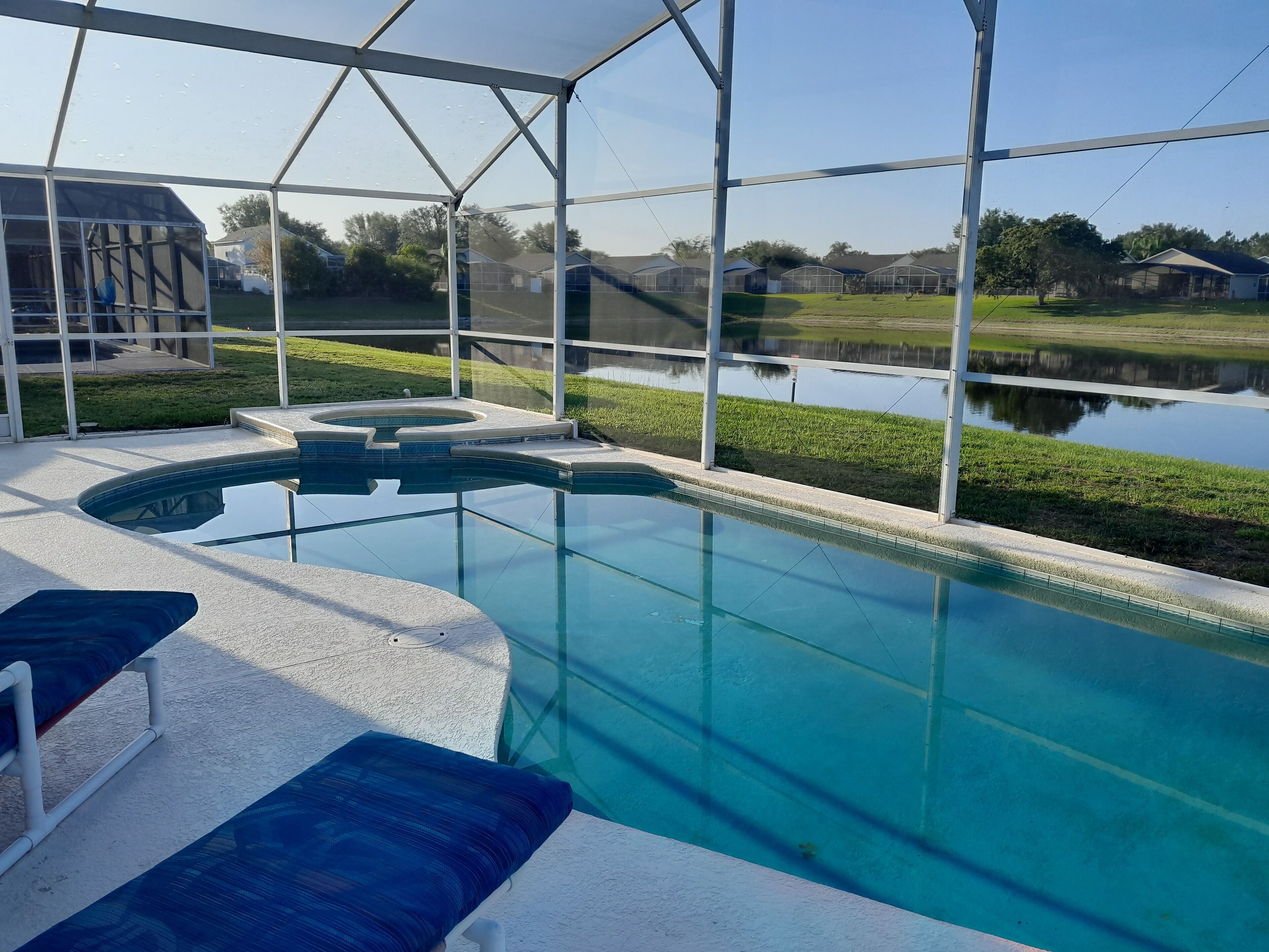 A swimming pool with a glass enclosure, blue cushioned lounge chairs, and a view of a lake and houses in the background.