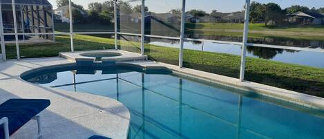 A swimming pool with a glass enclosure, blue cushioned lounge chairs, and a view of a lake and houses in the background.