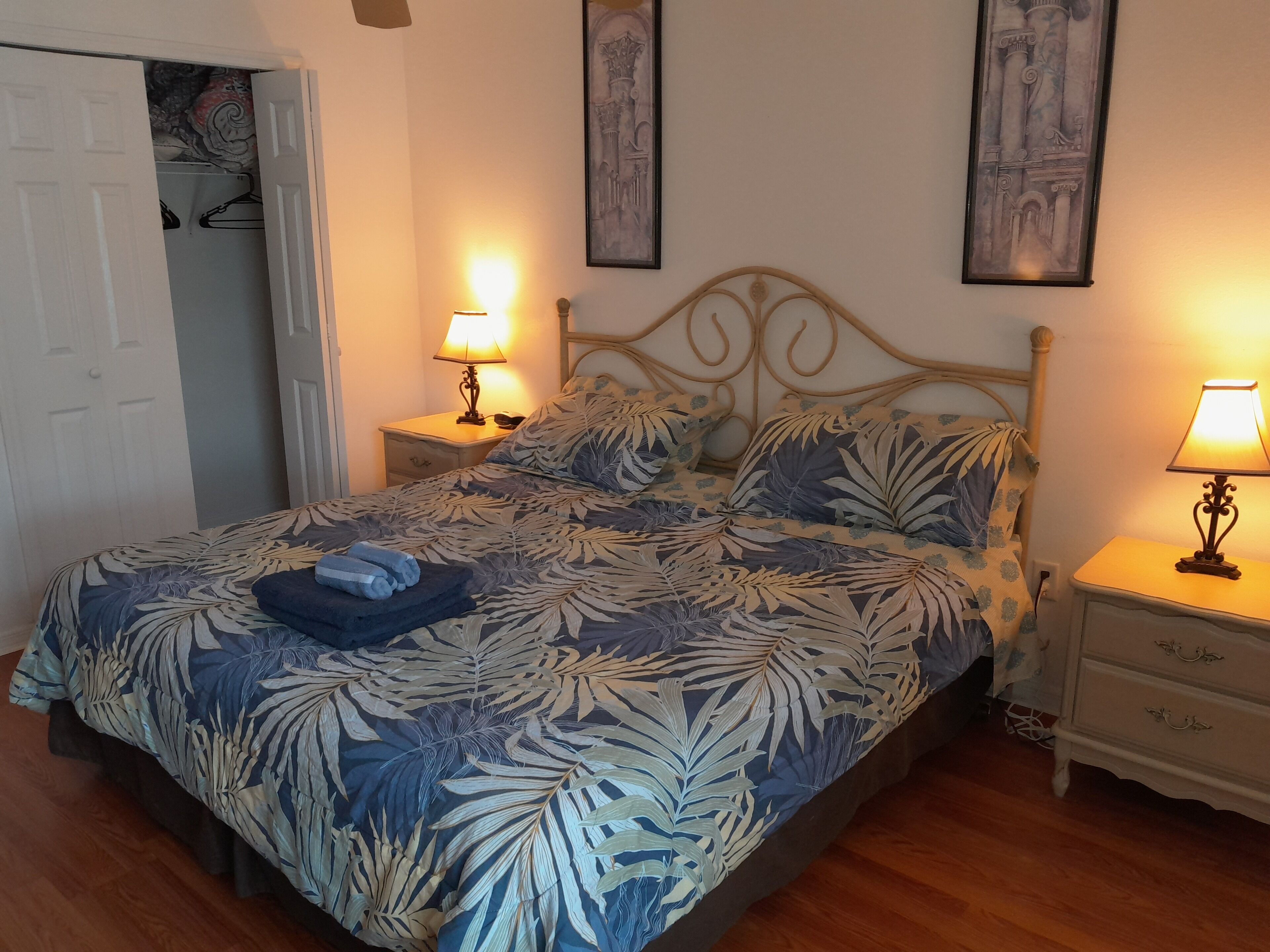 Un dormitorio con una cama, dos mesitas de noche con lámparas, una puerta y dos cuadros en la pared.