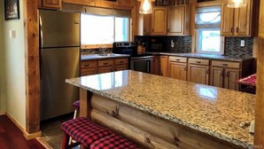 Una cocina con gabinetes de madera, encimeras de granito, un refrigerador de acero inoxidable e iluminación colgante.