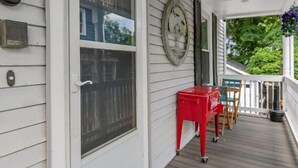 Een overdekt terras met een rode tafel en stoelen, een groot raam met jaloezieën en een klok aan de muur.