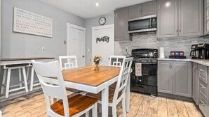 Een moderne keuken met grijze kastjes, een houten eettafel, witte stoelen, een klok en een bord met de tekst 'schilderijen' aan de muur.