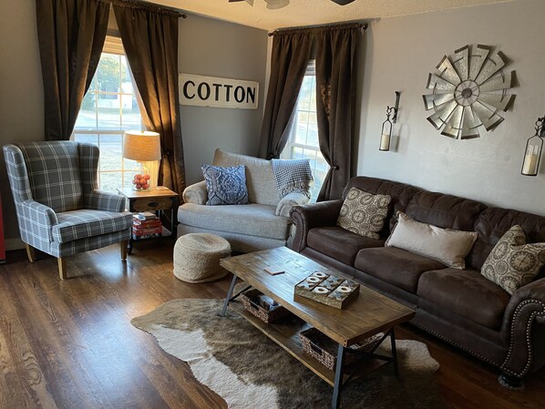 Un salon chaleureux avec un fauteuil à carreaux, un canapé gris et un divan en cuir marron. Sur le mur, on peut voir une enseigne indiquant « COTON », un moulin à vent décoratif et une table basse en bois sur laquelle est posé un jeu de société.