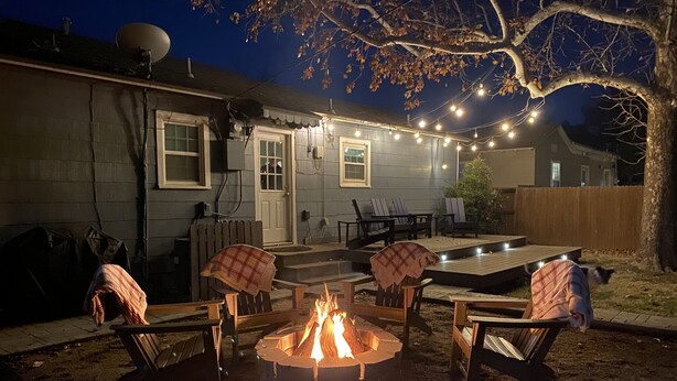 Un'area patio esterna con un braciere circondato da sedie e un tavolo, luci decorative sospese e un pavimento in legno.