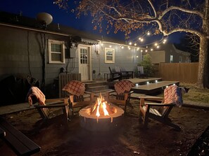 An outdoor patio area with a fire pit surrounded by chairs and a table, string lights overhead, and a wooden deck.