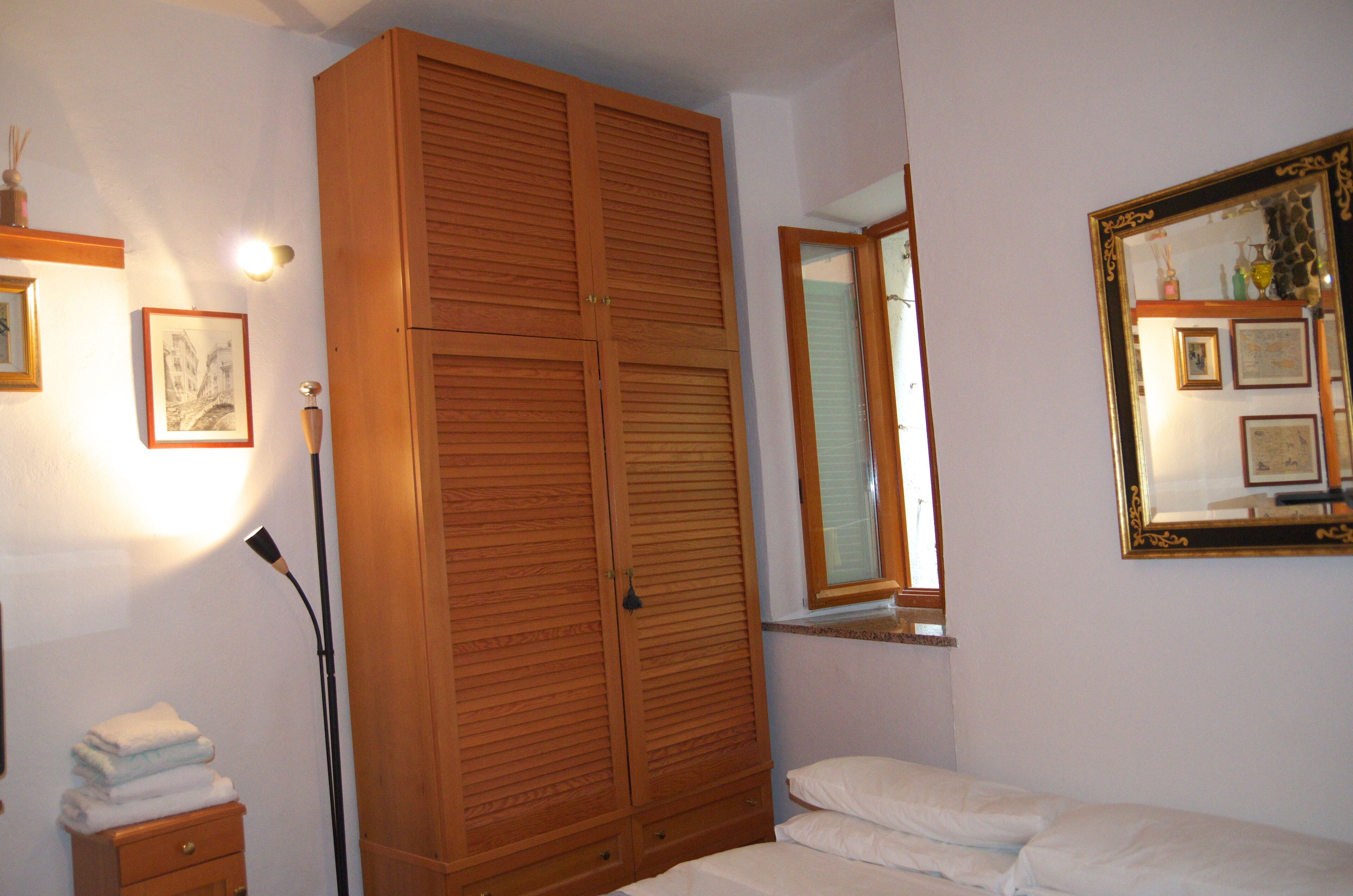 Une chambre avec une armoire en bois, une lampe de chevet, un lit avec une literie blanche et un miroir fixé au mur.