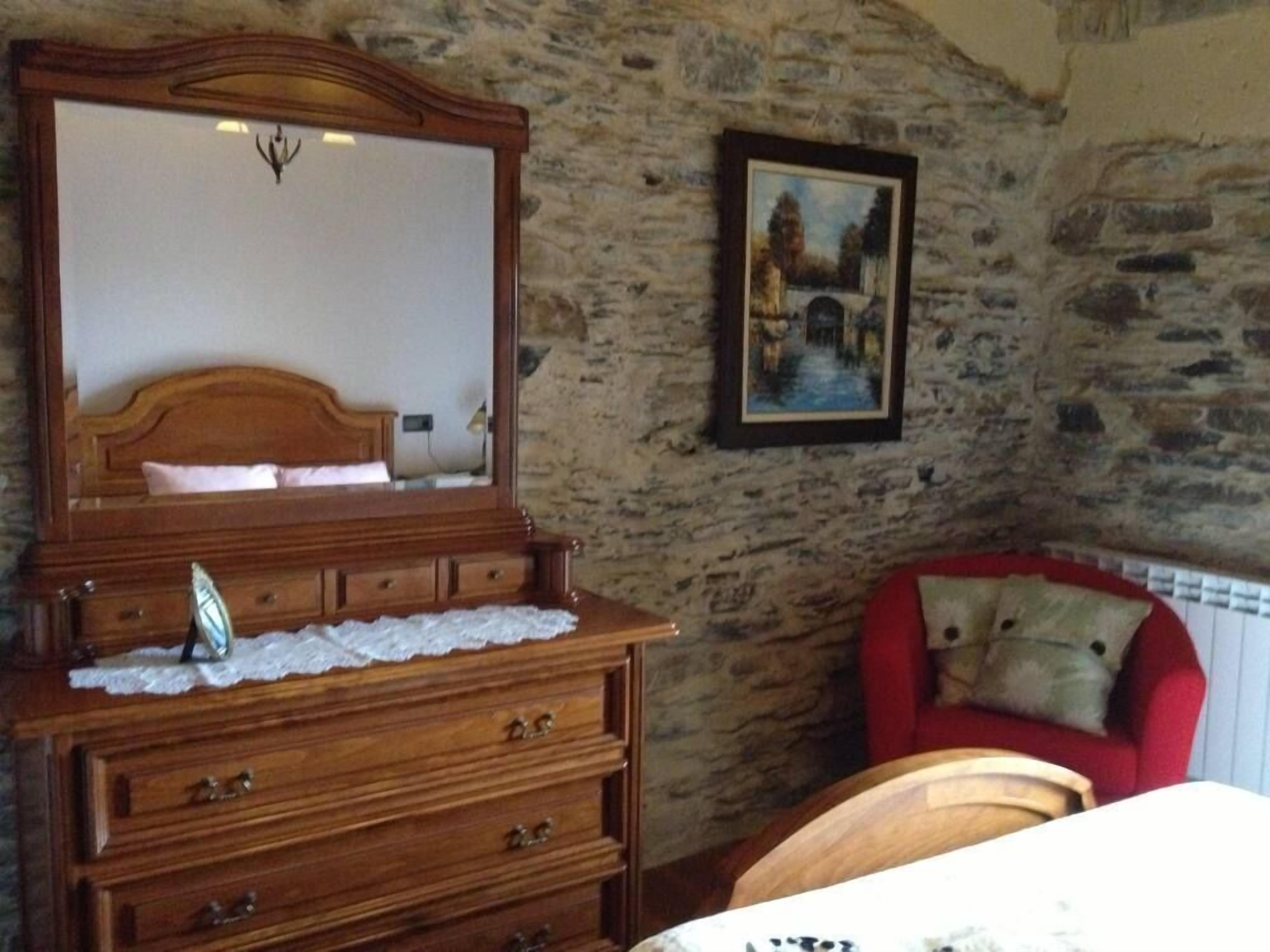 A bedroom with a wooden dresser, a mirror, a red chair, and a stone wall.
