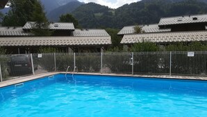 En swimmingpool med træterrasse, omgivet af et metalhegn, med bjerge i baggrunden.