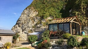 Una cabaña rústica con un porche acristalado, rodeada de un jardín con diversas plantas y vistas a una ladera rocosa.