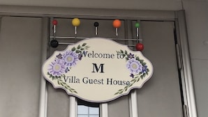 Une enseigne accueillante pour la Villa Guest House, ornée de motifs floraux et portant la lettre « M », qui indique le nom de l’établissement.