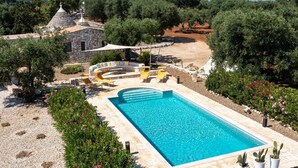 Una piscina rectangular con borde de piedra, rodeada por un jardín con diversas plantas y un pequeño edificio de piedra con techo cónico.