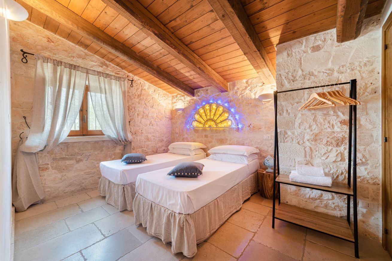 Une pièce confortable avec deux lits, un plafond en bois, des murs en pierre, une fenêtre avec des rideaux et une étagère fixée au mur où sont rangées des serviettes.
