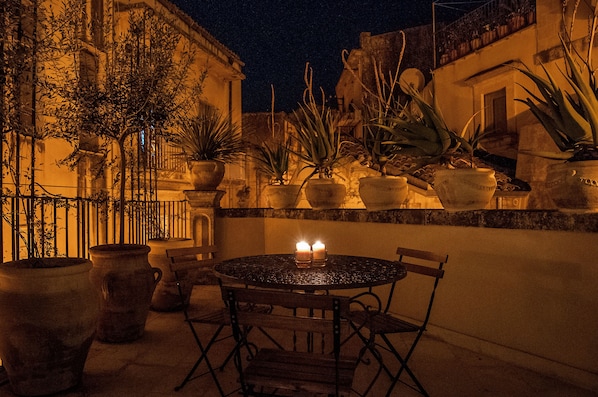 An outdoor patio with a round table and chairs, surrounded by large potted plants and a railing.