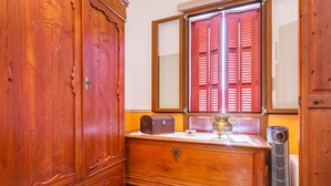 Une commode en bois avec un miroir au-dessus, un radiateur et une fenêtre avec des volets rouges.