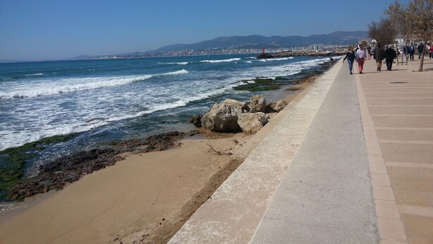 Une promenade en bord de mer, avec un chemin piétonnier en béton longeant la mer, des affleurements rocheux et une vue lointaine sur la ville.