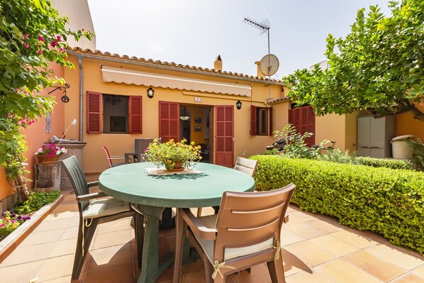 A patio with a round table and chairs, a potted plant, and a building with red shutters and a satellite dish.