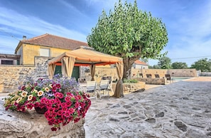 A stone-paved courtyard with a covered seating area, a large tree, and a flower bed with pink and purple flowers. - Holiday House Oliva - with a private pool and 10 min to the beach by MyWaycation (Podlug)
