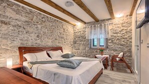 Un dormitorio con pared de piedra, vigas de madera en el techo, una cama con ropa blanca, una mesita de noche con lámpara y una ventana con cortinas.