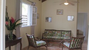 Un salon comprenant un canapé, deux fauteuils, une table avec un vase de fleurs, un ventilateur de plafond et une fenêtre avec des rideaux.